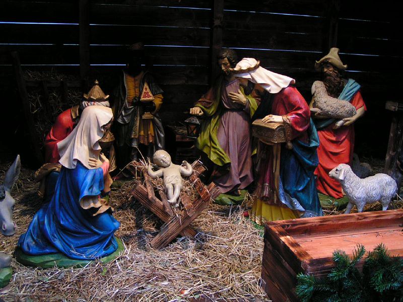 Foto van een kerststal met poppen. Op het stro zien we Maria in het blauw bij de kribbe waar kindje Jezus ligt, Joezef staat aan de andere kant en op de achtergrond de drie wijzen en een herder met lammetjes.