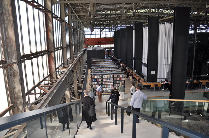 Foto van de LocHal in Tilburg. Mensen lopen van een betonnen trap naar beneden waar rijen boekenkasten staan en mensen aan smalle tafels zitten te werken. Industrieel gebouw.