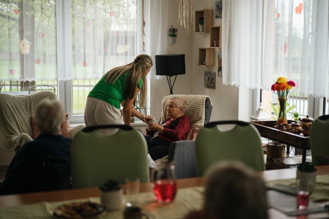 Foto van de gemeenschappelijke ruimte in verpleeghuizen. Een verzorger geeft een kopje drinken aan een van de ouderen die op de stoel zit in de ruimte.